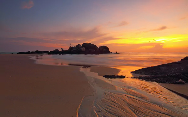 A serene seascape at sunrise, featuring an orange glow over the ocean, gently lapping waves on the sand, and rocky formations on the horizon. An inspiring natural backdrop.