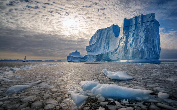 HD desktop wallpaper featuring an iceberg floating in the ocean under a cloudy sky with a distant sailboat, showcasing the serene beauty of ice and nature.