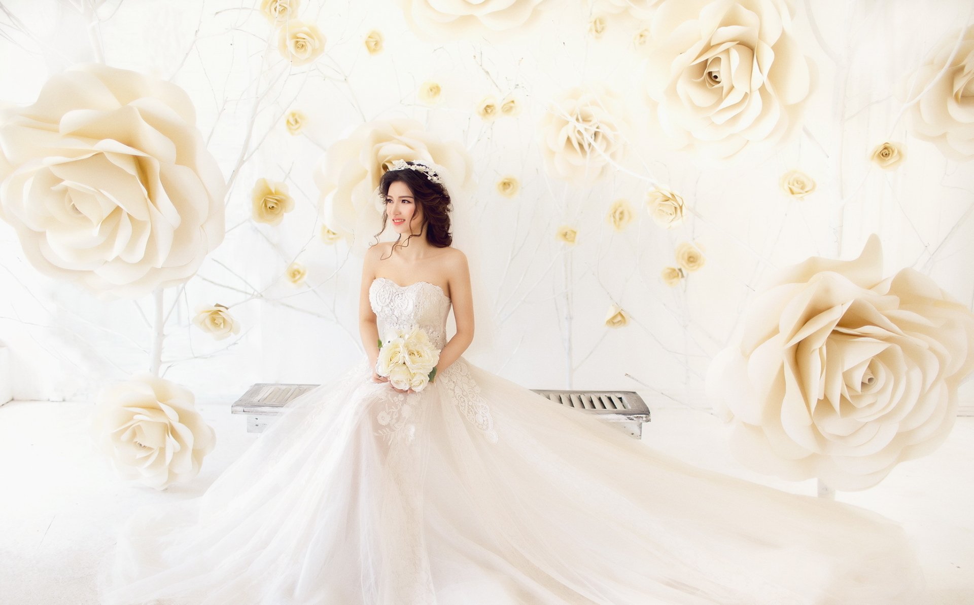 HD PC desktop wallpaper of a brunette bride in a white wedding dress holding a bouquet, framed by oversized white flowers, her gown flowing across the floor.