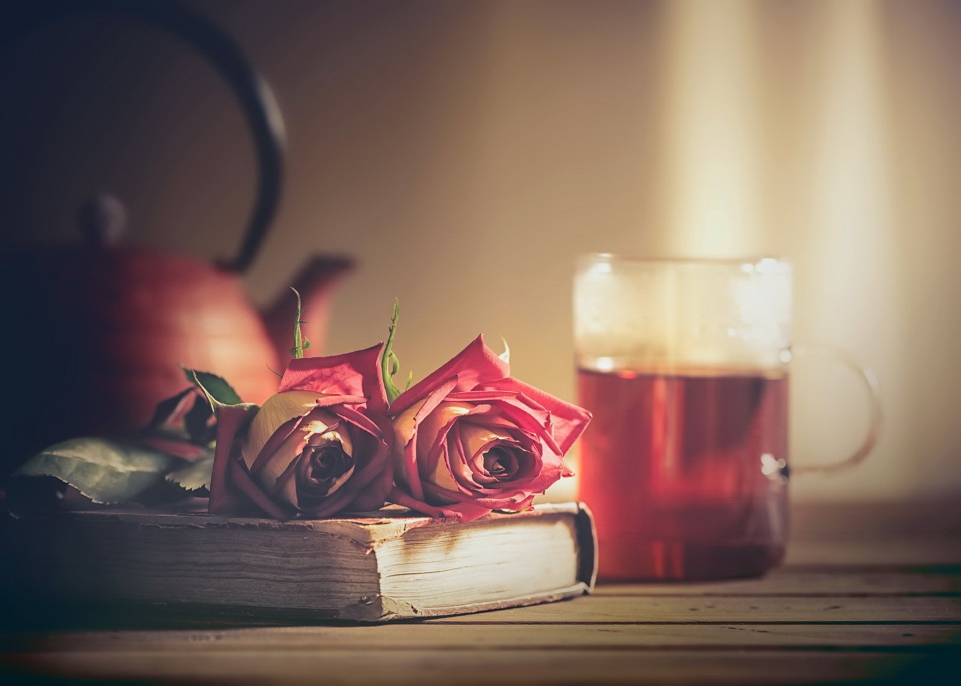 A still life HD desktop wallpaper featuring a blurred teapot, two pink roses resting on a book, and a glass mug of tea on a wooden surface.