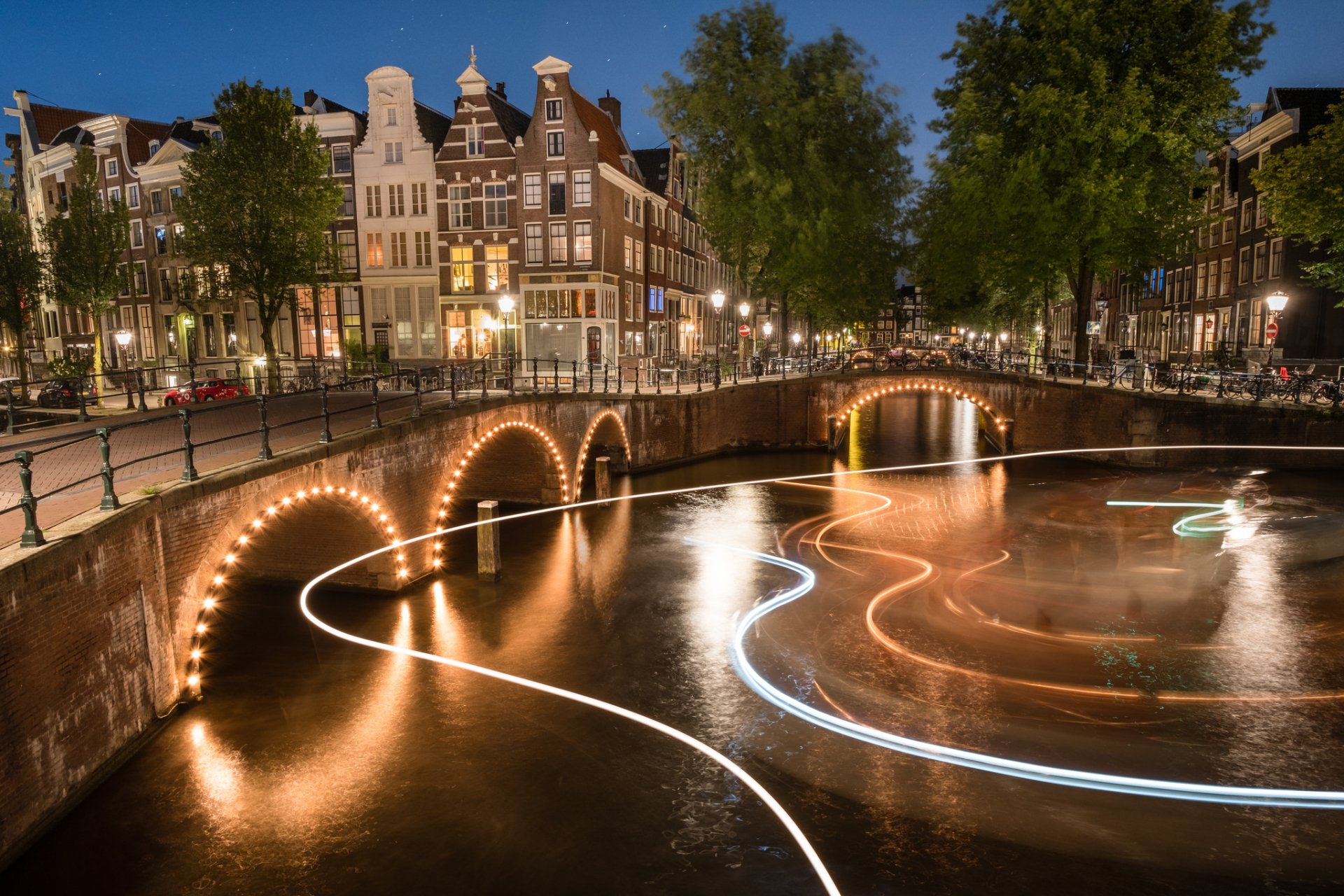 HD desktop wallpaper: night time-lapse light trails over an Amsterdam canal, illuminated arched man-made bridges and historic Dutch city buildings in the Netherlands.