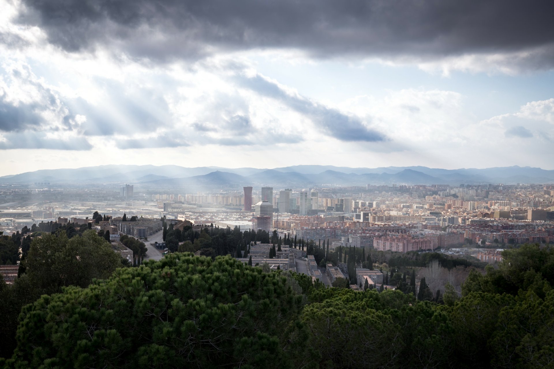 Sunbeam filters through clouds over the cityscape of Barcelona, Spain, showcasing a vast man-made urban landscape with mountains in the distance.
