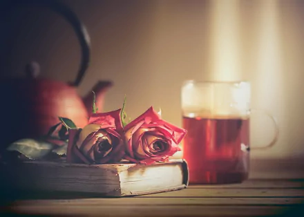 A still life HD desktop wallpaper featuring a blurred teapot, two pink roses resting on a book, and a glass mug of tea on a wooden surface.