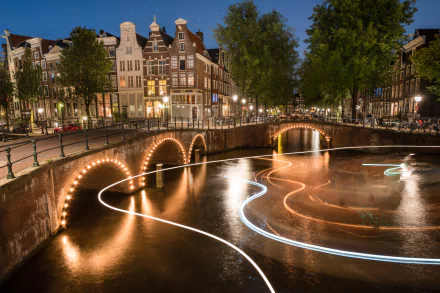 HD desktop wallpaper: night time-lapse light trails over an Amsterdam canal, illuminated arched man-made bridges and historic Dutch city buildings in the Netherlands.