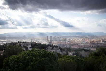 Sunbeam filters through clouds over the cityscape of Barcelona, Spain, showcasing a vast man-made urban landscape with mountains in the distance.
