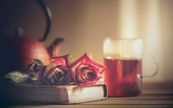 A still life HD desktop wallpaper featuring a blurred teapot, two pink roses resting on a book, and a glass mug of tea on a wooden surface.