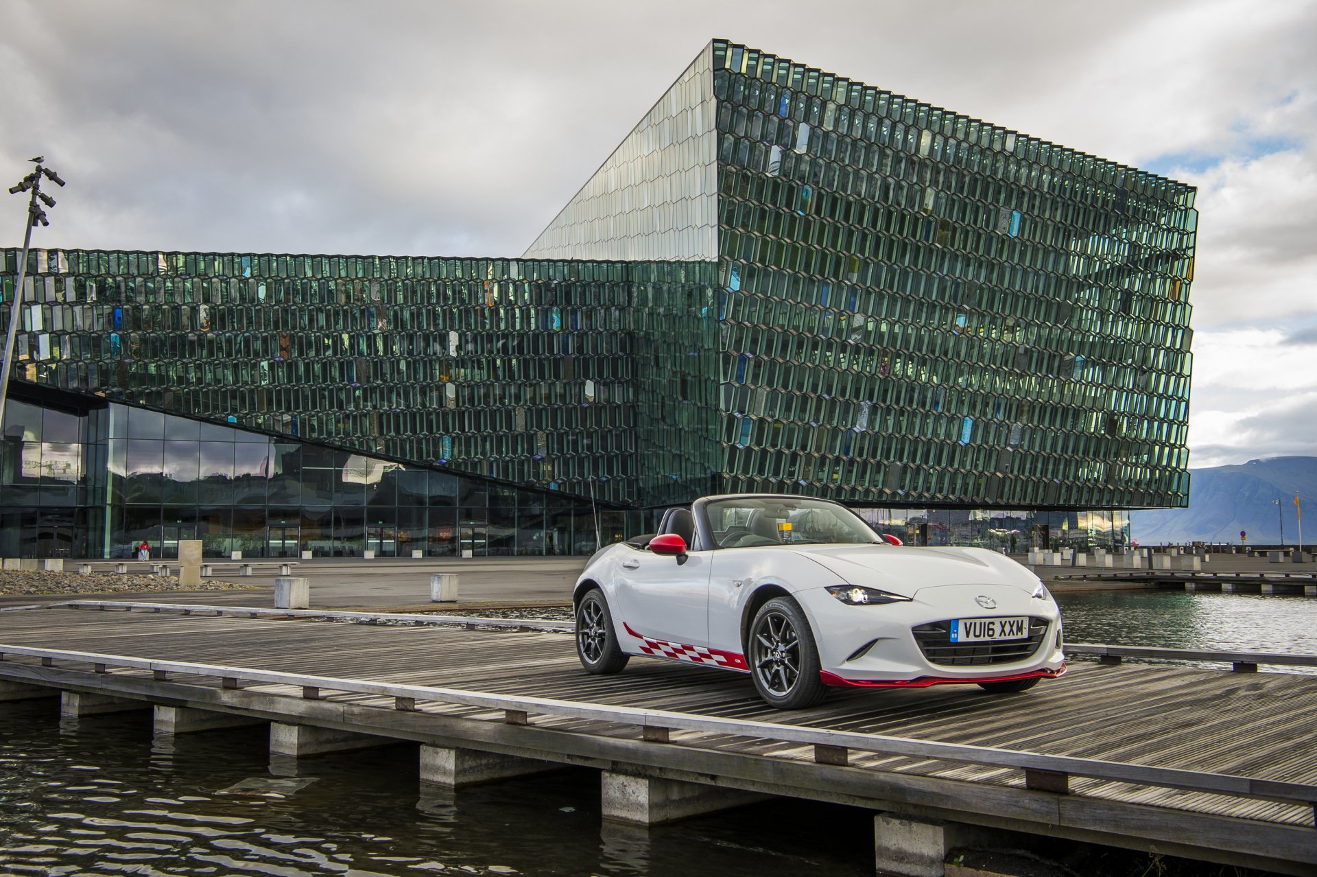 A white Mazda MX-5 sports car parked on a wooden dock with a modern glass building and cloudy sky in the background.
