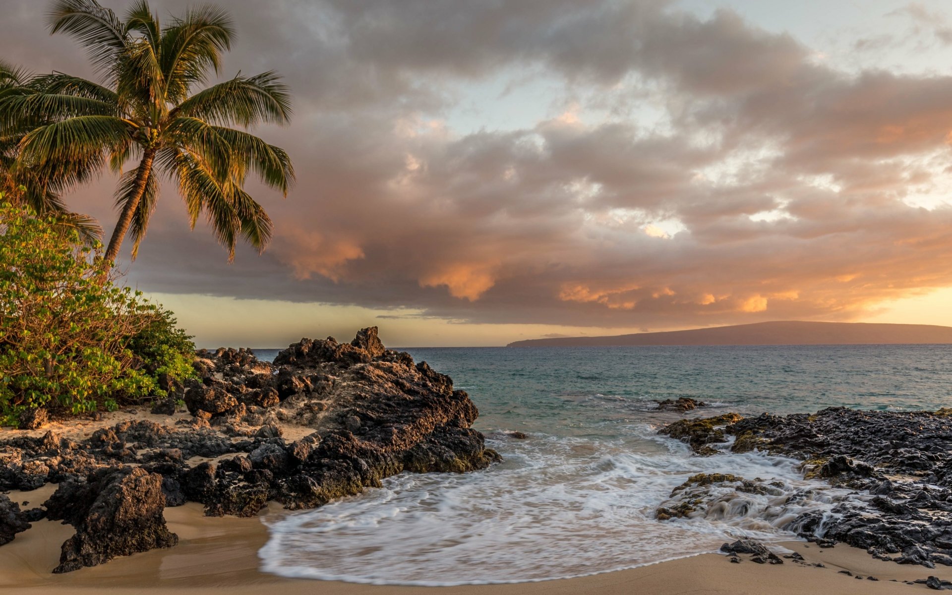 HD desktop wallpaper showcasing a serene Hawaiian beach with palm trees, rocky shores, ocean waves, and dramatic clouds at sunset.