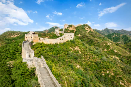 The Great Wall of China winds through a lush landscape, showcasing its majestic man-made structure against a backdrop of rolling hills and vibrant blue skies.