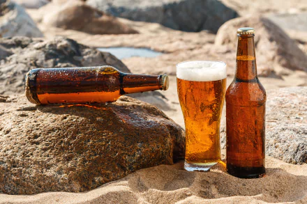 A sunny beach scene featuring a glass of beer alongside two brown bottles on a sandy surface, with rocks in the background, captured in vibrant 4K Ultra HD.