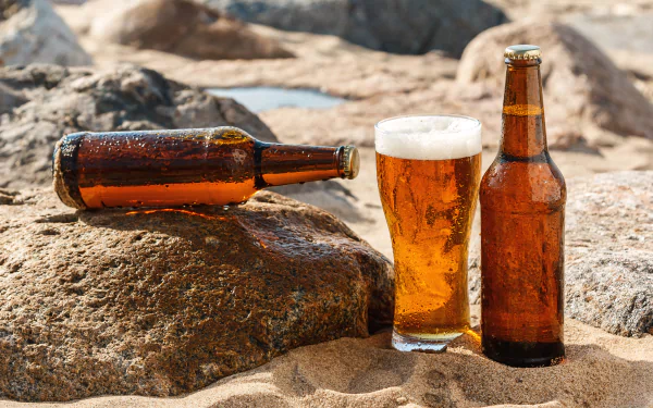 A sunny beach scene featuring a glass of beer alongside two brown bottles on a sandy surface, with rocks in the background, captured in vibrant 4K Ultra HD.