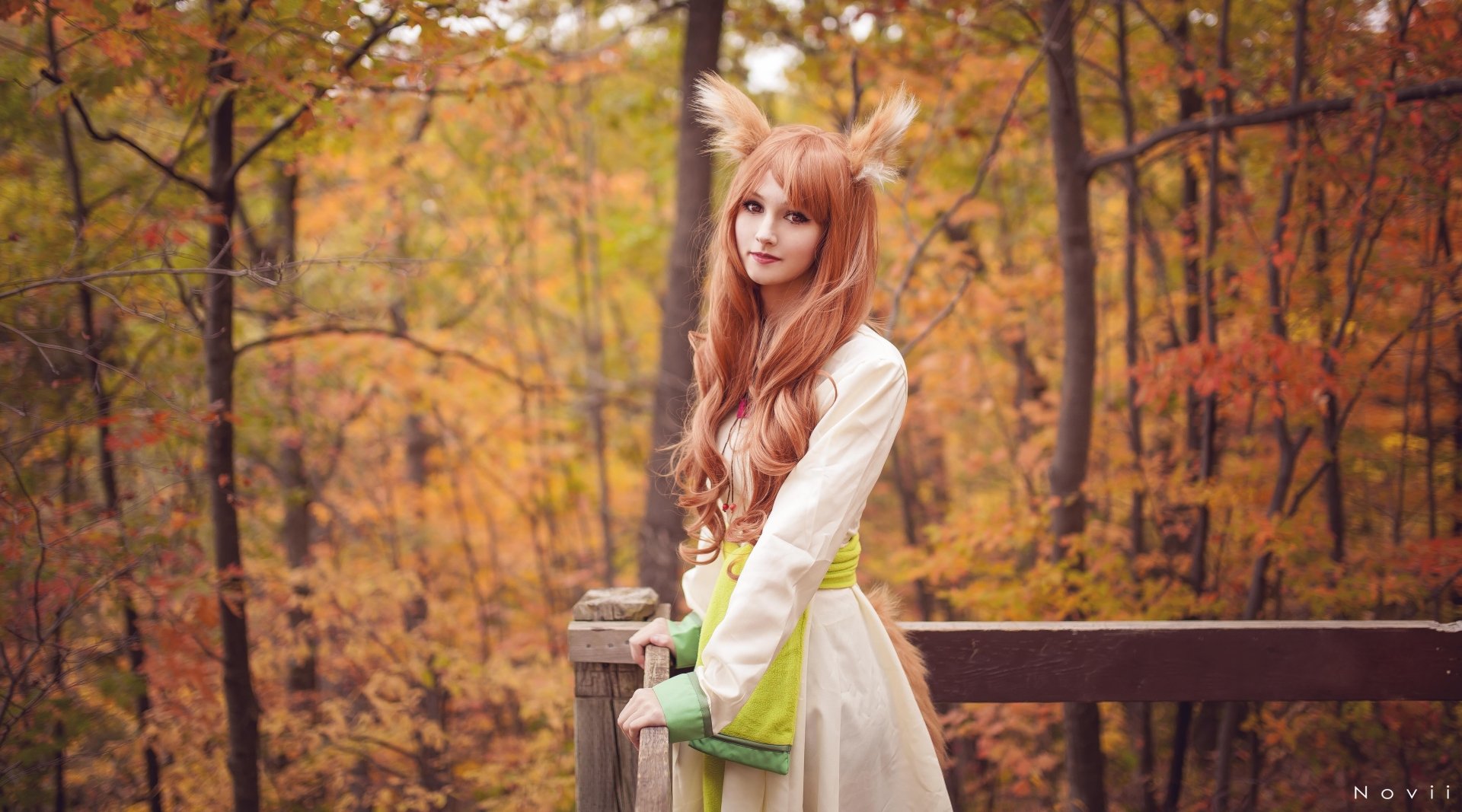 A woman cosplaying as Holo from Spice & Wolf stands in a vibrant autumn setting, embodying the character's spirit amidst colorful foliage, creating a captivating HD wallpaper.