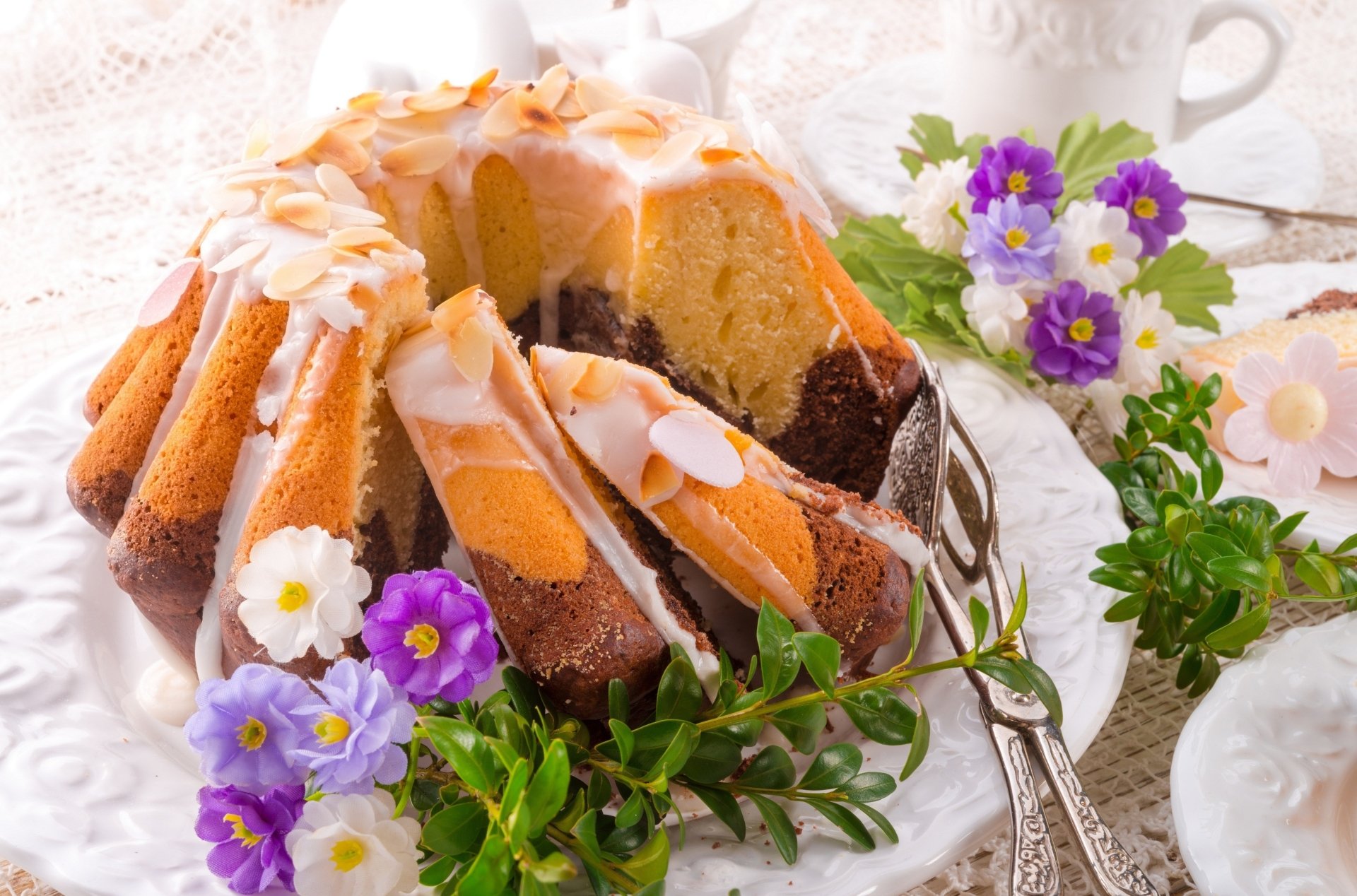 A 4K Ultra HD wallpaper of a glazed marble bundt cake decorated with almond slices, surrounded by purple and white flowers on a lace tablecloth.