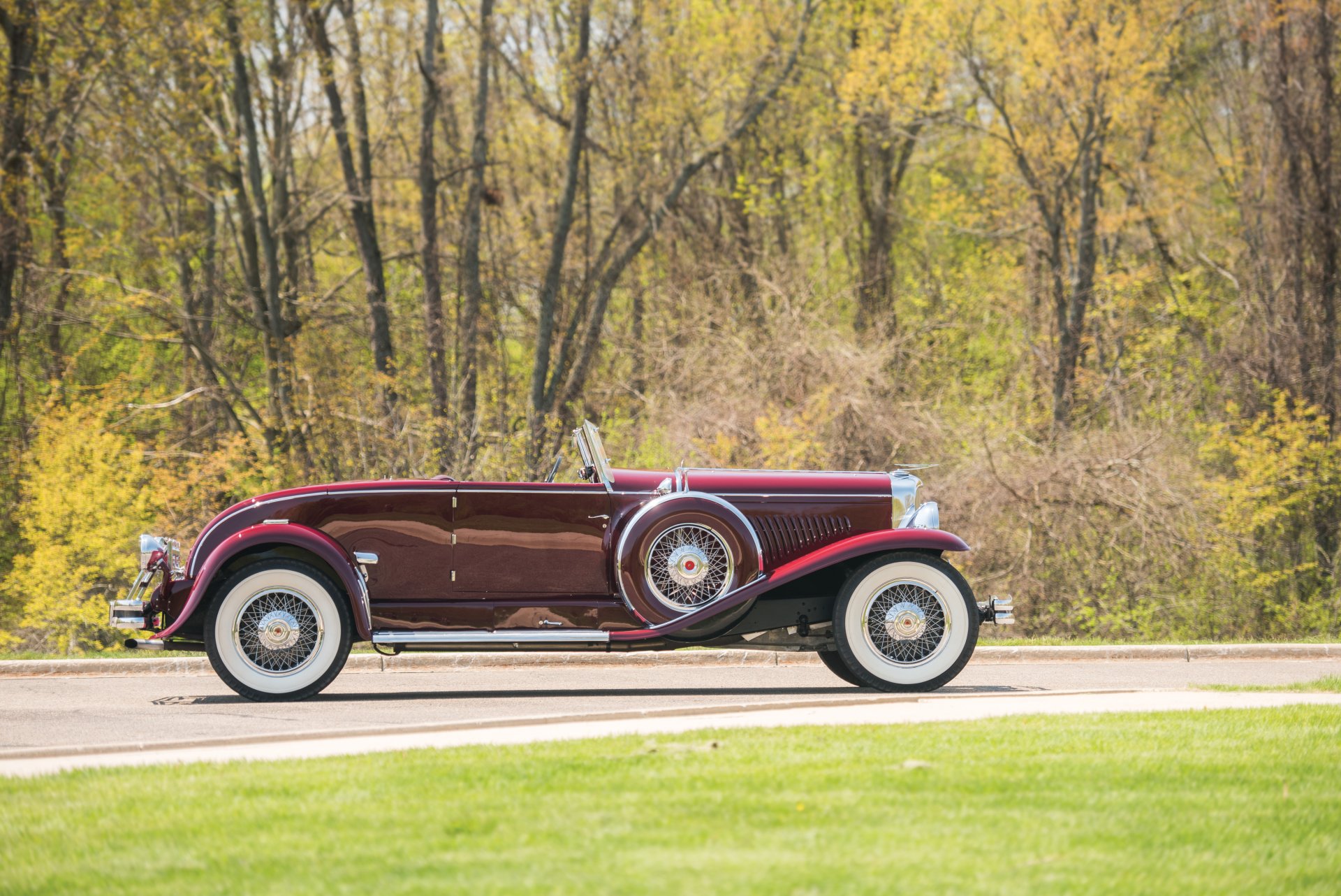 1929 Duesenberg Model J Disappearing Top Convertible Coupe parked on a road with autumn trees in the background, captured as an HD vintage car desktop wallpaper.
