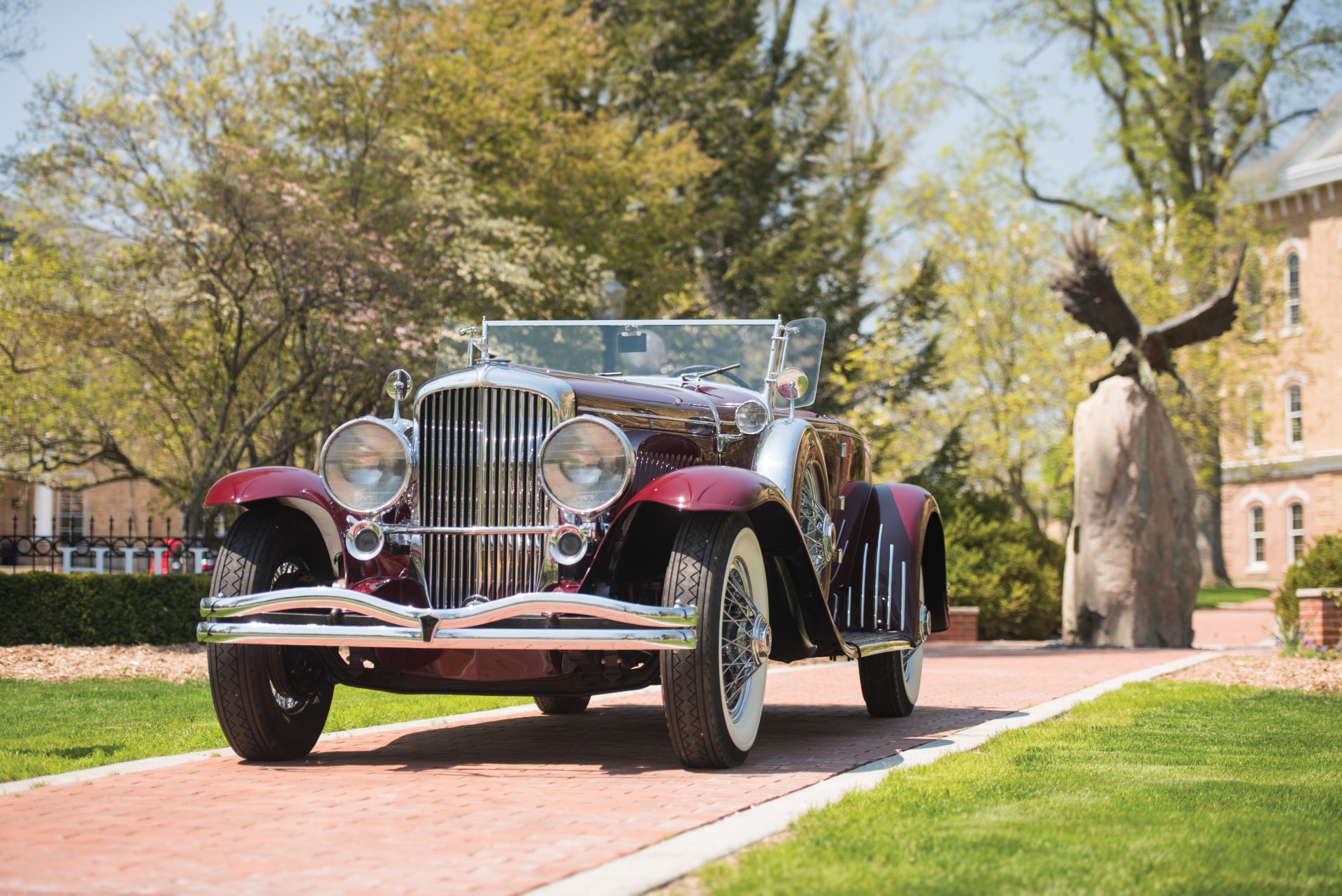 Vintage 1929 Duesenberg Model J Disappearing Top Convertible Coupe parked on a brick path with trees and a large eagle sculpture in the background.