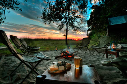 A cozy camping scene at sunset features a tree, a bonfire, chairs, and a table with beer and food, all illuminated by lanterns, creating a warm outdoor atmosphere.