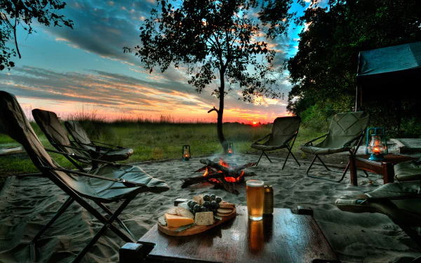 A cozy camping scene at sunset features a tree, a bonfire, chairs, and a table with beer and food, all illuminated by lanterns, creating a warm outdoor atmosphere.
