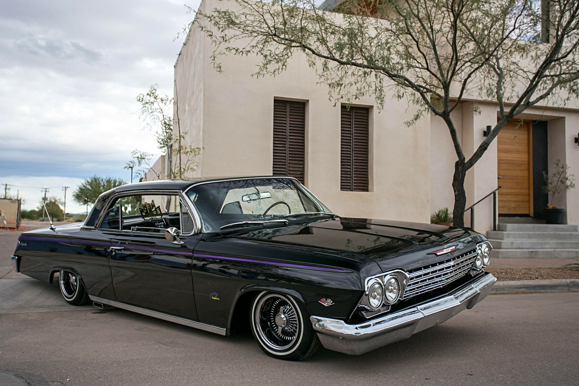 HD desktop wallpaper featuring a classic black Chevrolet Impala lowrider muscle car parked on a street with a modern building and tree in the background.
