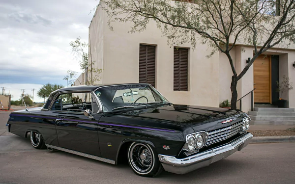HD desktop wallpaper featuring a classic black Chevrolet Impala lowrider muscle car parked on a street with a modern building and tree in the background.
