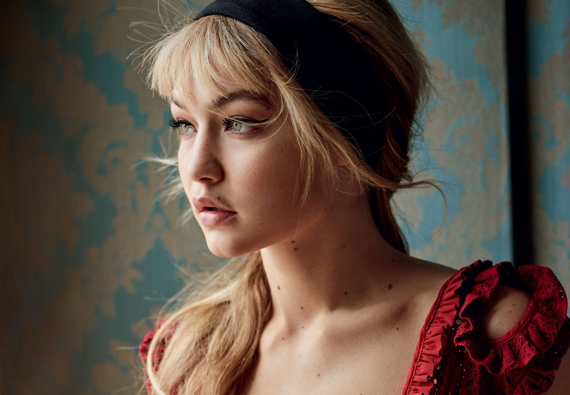 A close-up of blonde model Gigi Hadid, captured in soft lighting with a contemplative expression, wearing a red dress against a decorative background. High-definition wallpaper image.