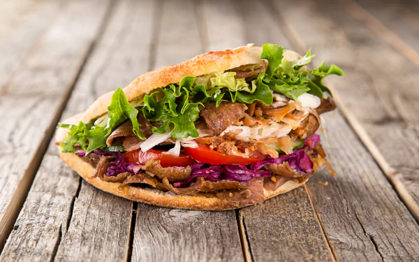 A close-up of a vibrant doner kebab filled with grilled meat, fresh lettuce, tomatoes, and onions, set on rustic wooden planks. 4K Ultra HD PC desktop wallpaper.