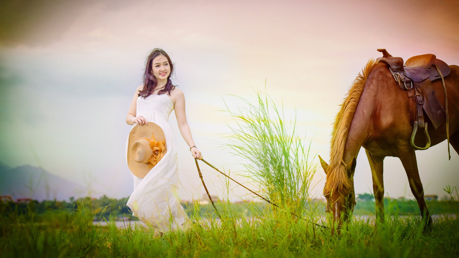 A brunette woman in a white dress and hat smiles while walking her horse through lush green grass, creating a serene and picturesque scene.
