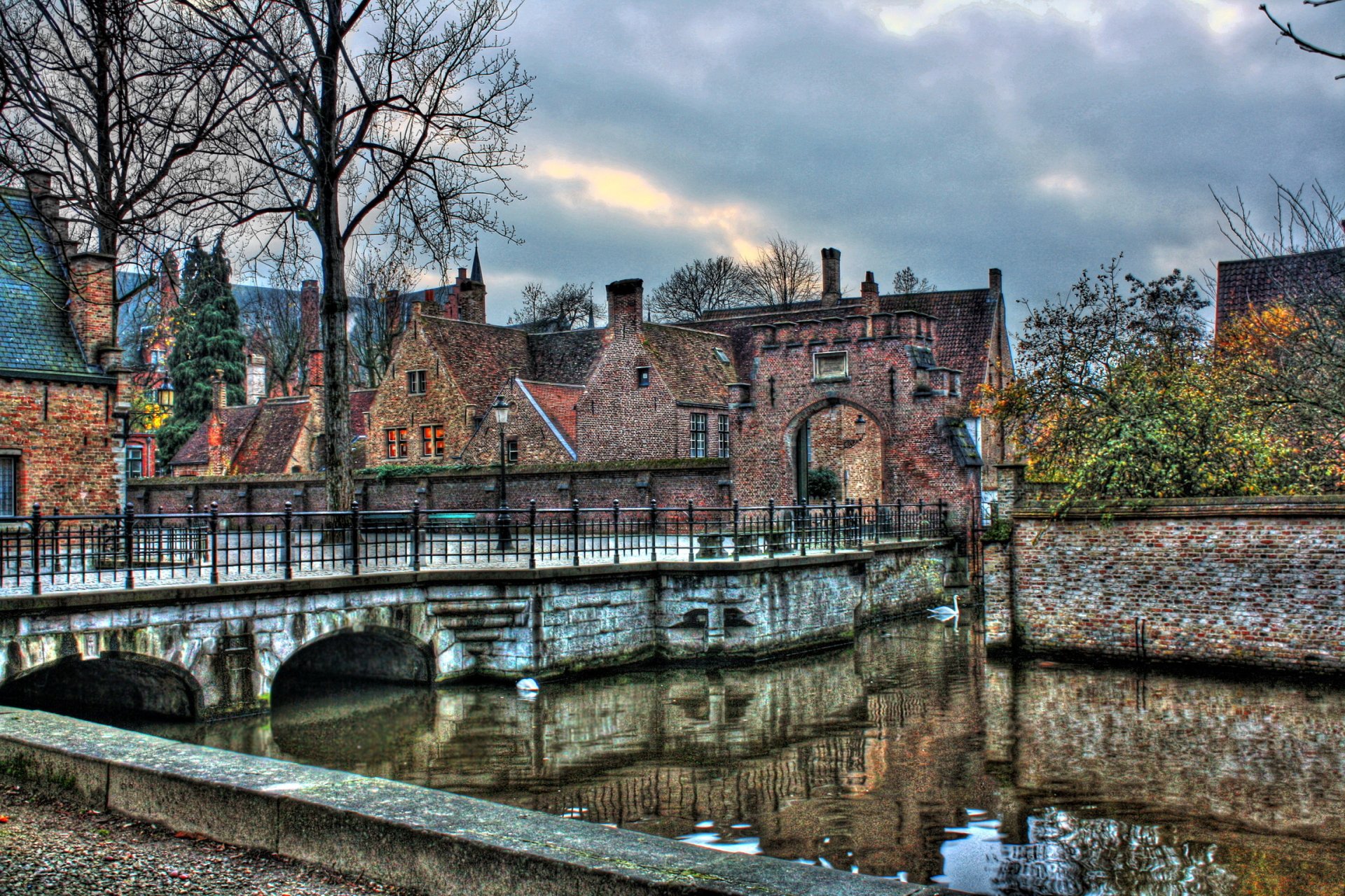 Scenic view of a man-made stone bridge over a river in the town of Bruges, Belgium, with historic houses lining the waterfront under a cloudy sky.