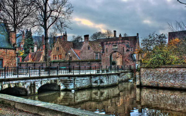 Scenic view of a man-made stone bridge over a river in the town of Bruges, Belgium, with historic houses lining the waterfront under a cloudy sky.