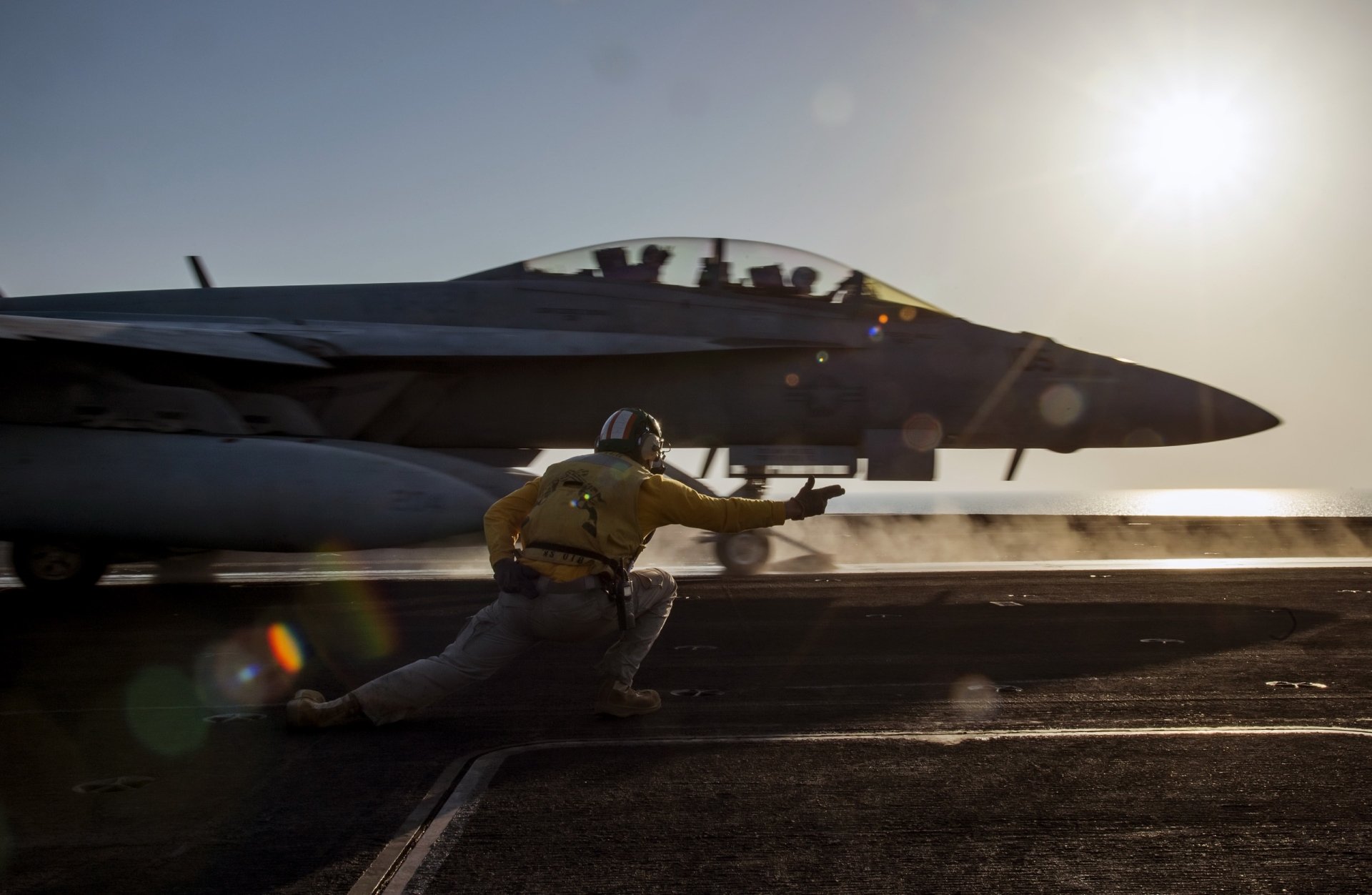 A Boeing F/A-18E/F Super Hornet warplane prepares for takeoff from an aircraft carrier deck at sunset, captured in a high-definition military jet fighter wallpaper.