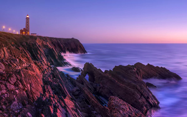 8K Ultra HD PC desktop wallpaper: dawn over the ocean horizon, rugged coast with rocky outcrops and a man-made lighthouse atop cliffs, soft pastel sky and long-exposure sea.