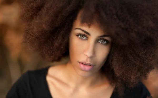 HD desktop wallpaper featuring a brunette model with hazel eyes and natural curly hair, showcasing a close-up of her face against a warm, blurred background.