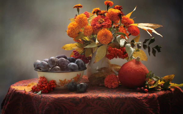 Still life HD wallpaper featuring vibrant orange marigold flowers, a small pumpkin, and a bowl of plums arranged on a richly textured tablecloth, evoking the colors of fall.