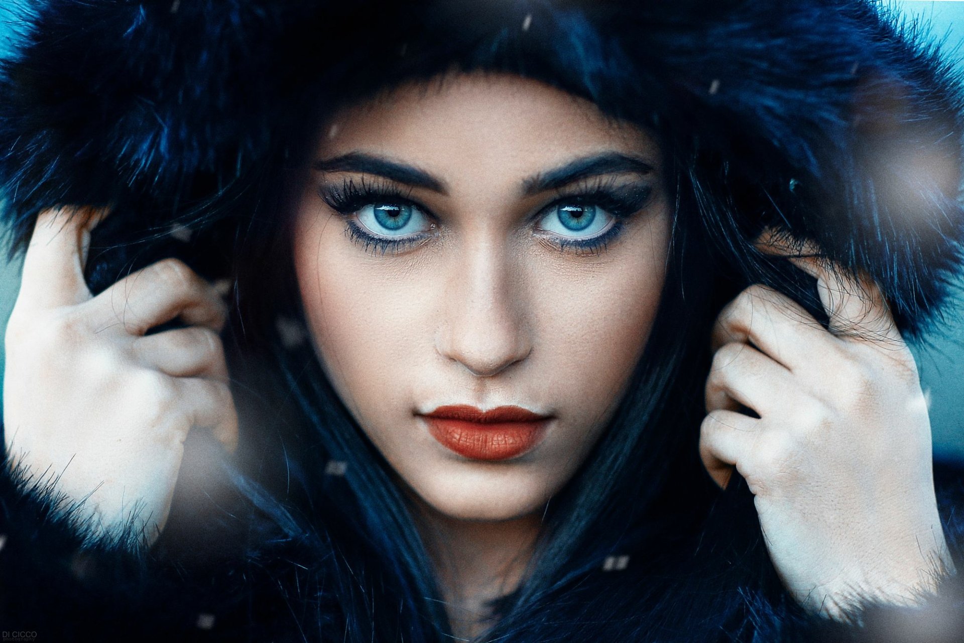 A striking model with blue eyes and bold red lipstick poses while wearing a fur-lined hood. This HD image captures her captivating gaze and features, making it an engaging desktop wallpaper.