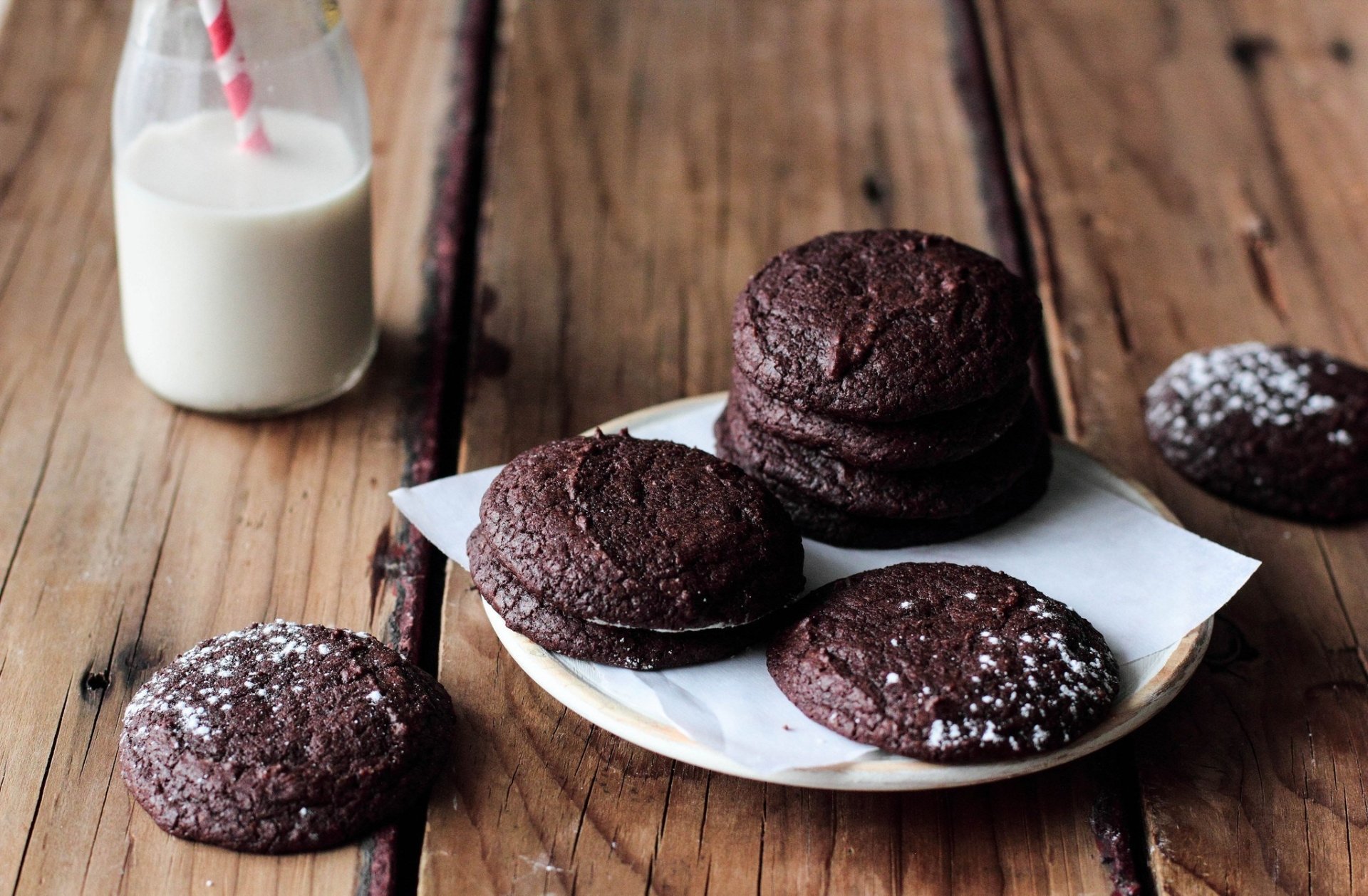 HD desktop wallpaper showing chocolate cookies on a plate with powdered sugar and a glass of milk with a straw on a wooden surface.