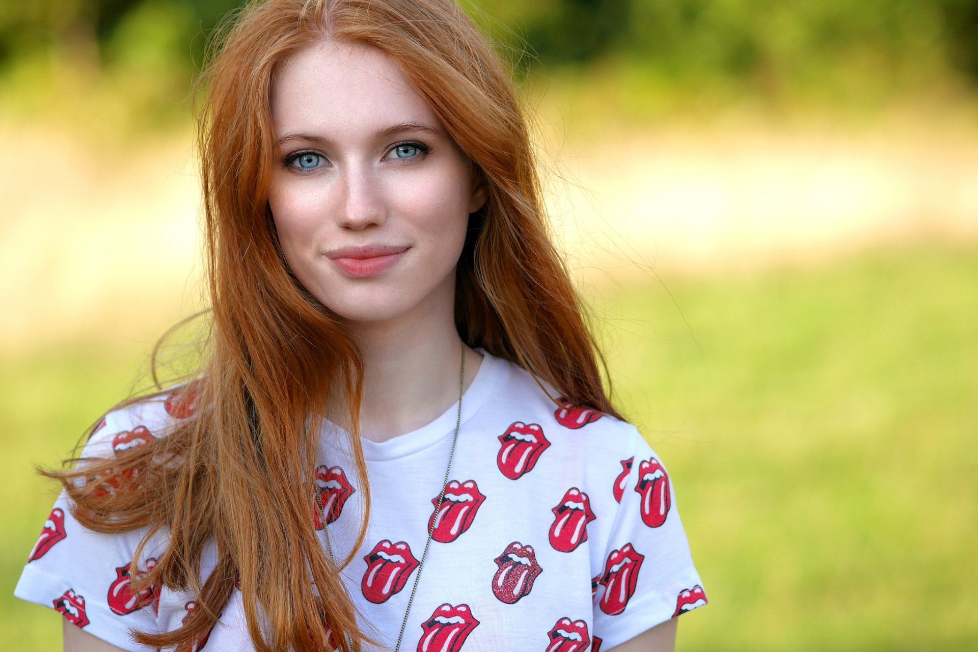 A smiling redhead woman with blue eyes wears a Rolling Stones shirt against a blurred green background, capturing a vibrant moment. High-definition desktop wallpaper.