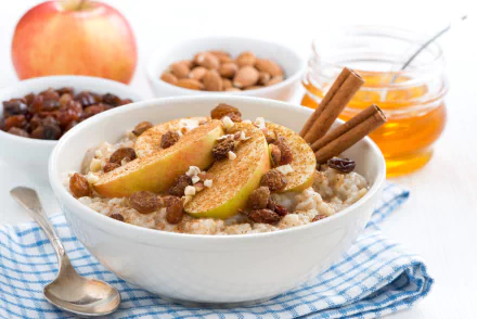 A bowl of porridge topped with apple slices, nuts, cinnamon sticks, and honey, surrounded by ingredients like apple, almonds, and honey jar on a white surface.