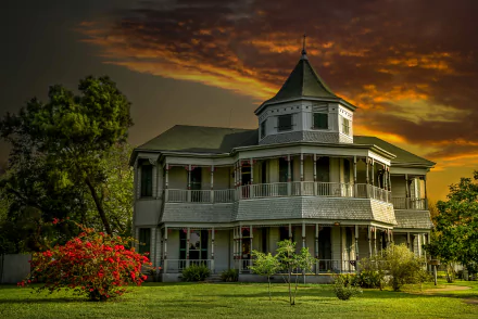 Victorian house surrounded by bushes and trees under a vibrant orange sunset sky, captured in an HD desktop wallpaper.