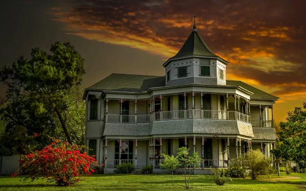 Victorian house surrounded by bushes and trees under a vibrant orange sunset sky, captured in an HD desktop wallpaper.
