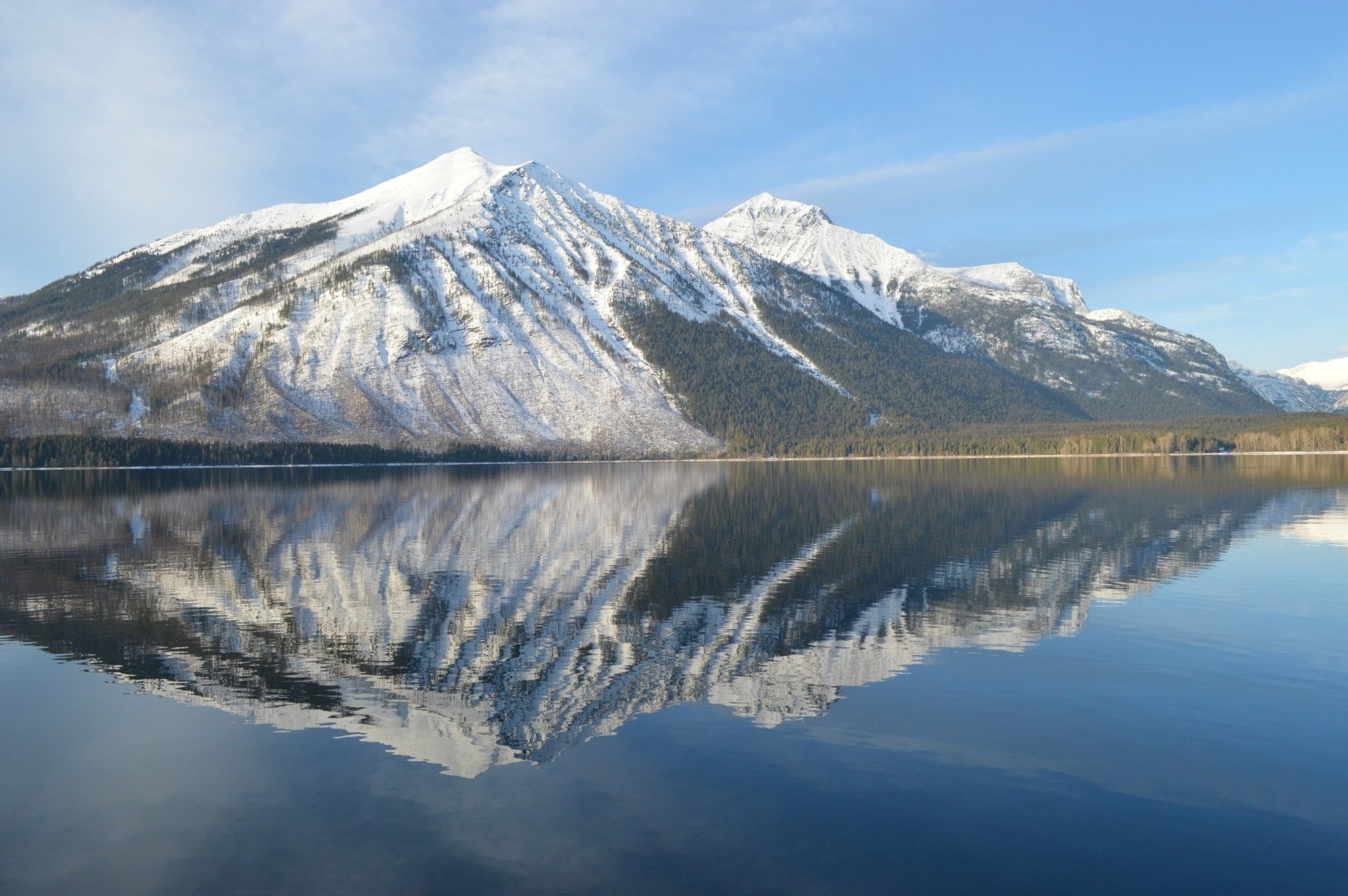 Snow-covered mountains of Glacier National Park in Montana reflect clearly on the calm waters of Lake McDonald under a bright blue sky.