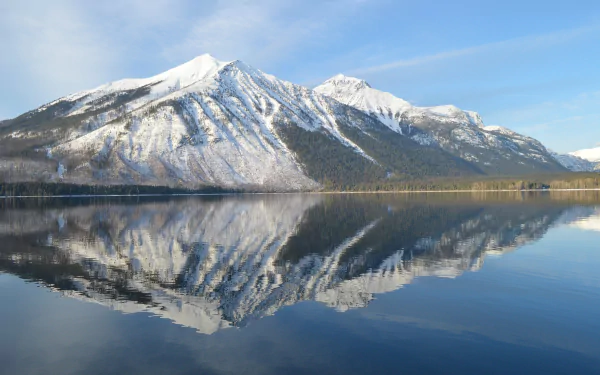 Snow-covered mountains of Glacier National Park in Montana reflect clearly on the calm waters of Lake McDonald under a bright blue sky.