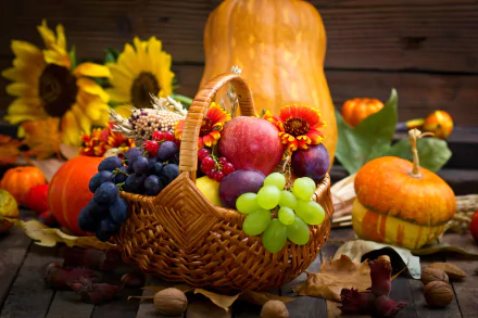 HD desktop wallpaper of a still life featuring a wicker basket filled with grapes, apples, gourds, and surrounded by walnuts, sunflower blossoms, and autumn leaves.