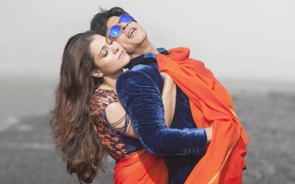 HD PC desktop wallpaper from the movie Dilwale — a romantic couple embraces on a muted shore; man in blue velvet jacket and sunglasses wrapped in an orange drape with the woman.