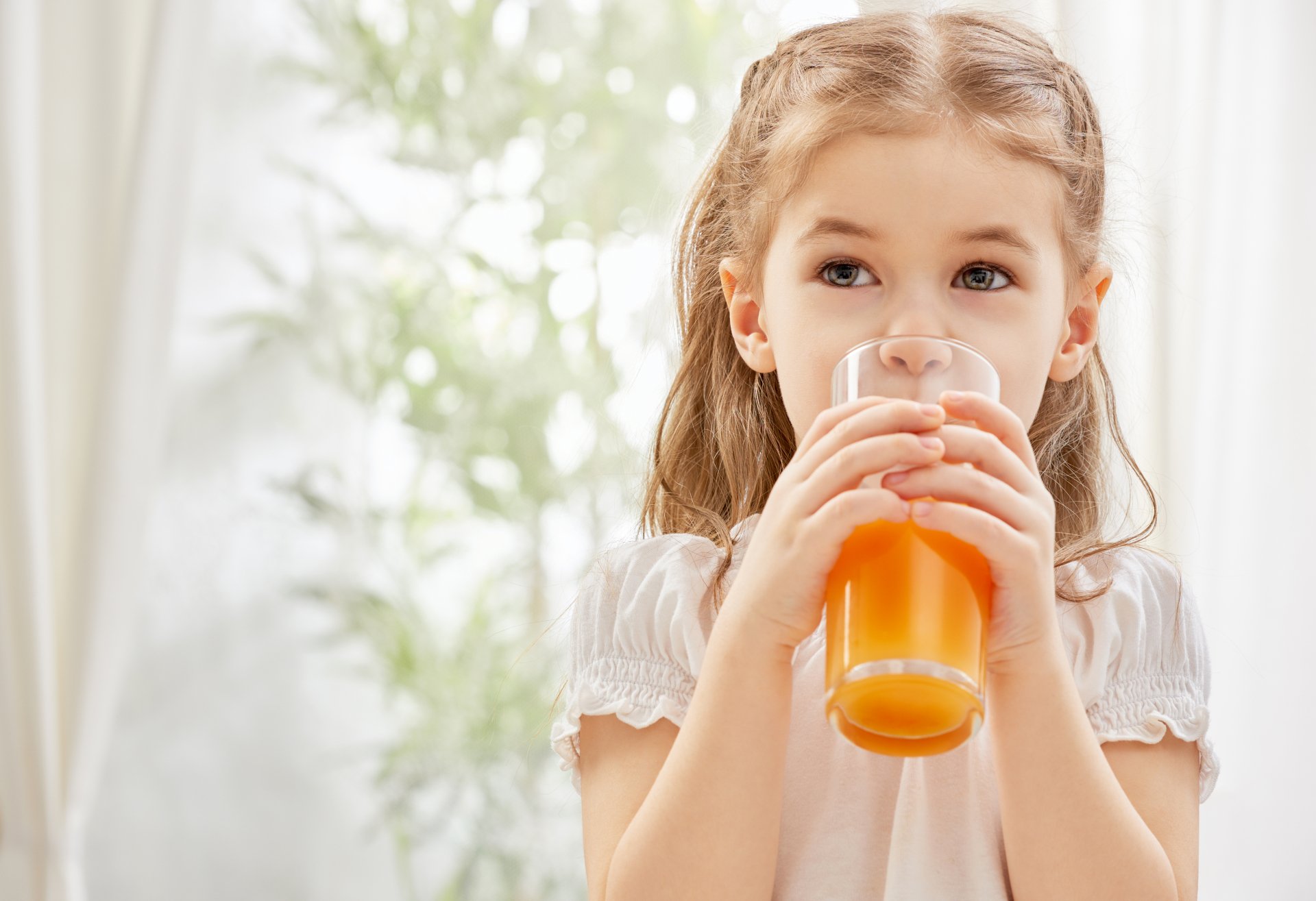 Blonde child with hazel eyes drinks orange juice in bright, softly lit setting, captured in 4K Ultra HD quality.