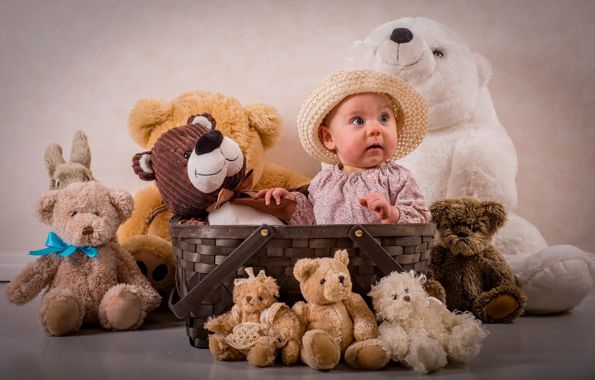 A baby wearing a hat sits in a basket surrounded by numerous teddy bears and stuffed animals, creating an adorable HD desktop wallpaper and background.
