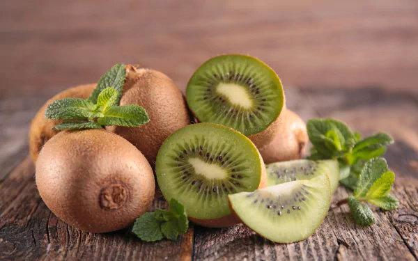 Close-up of fresh kiwi fruit, whole and sliced, accompanied by mint leaves on rustic wood, captured in vibrant detail as a 4K Ultra HD PC desktop wallpaper and background.