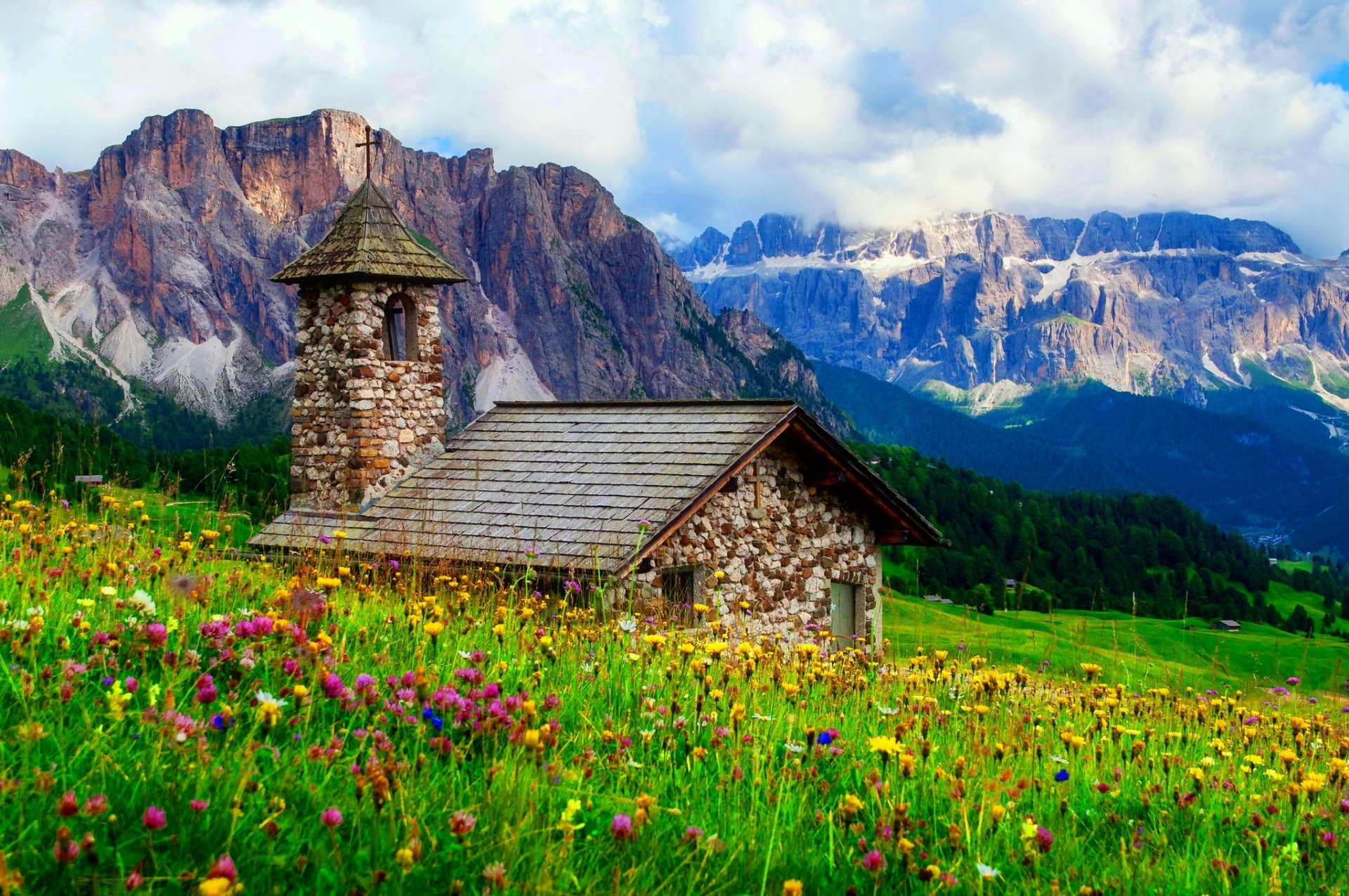 A stone chapel amidst a colorful flower field with towering mountains in the background under a partly cloudy sky, captured in a vibrant HD desktop wallpaper.