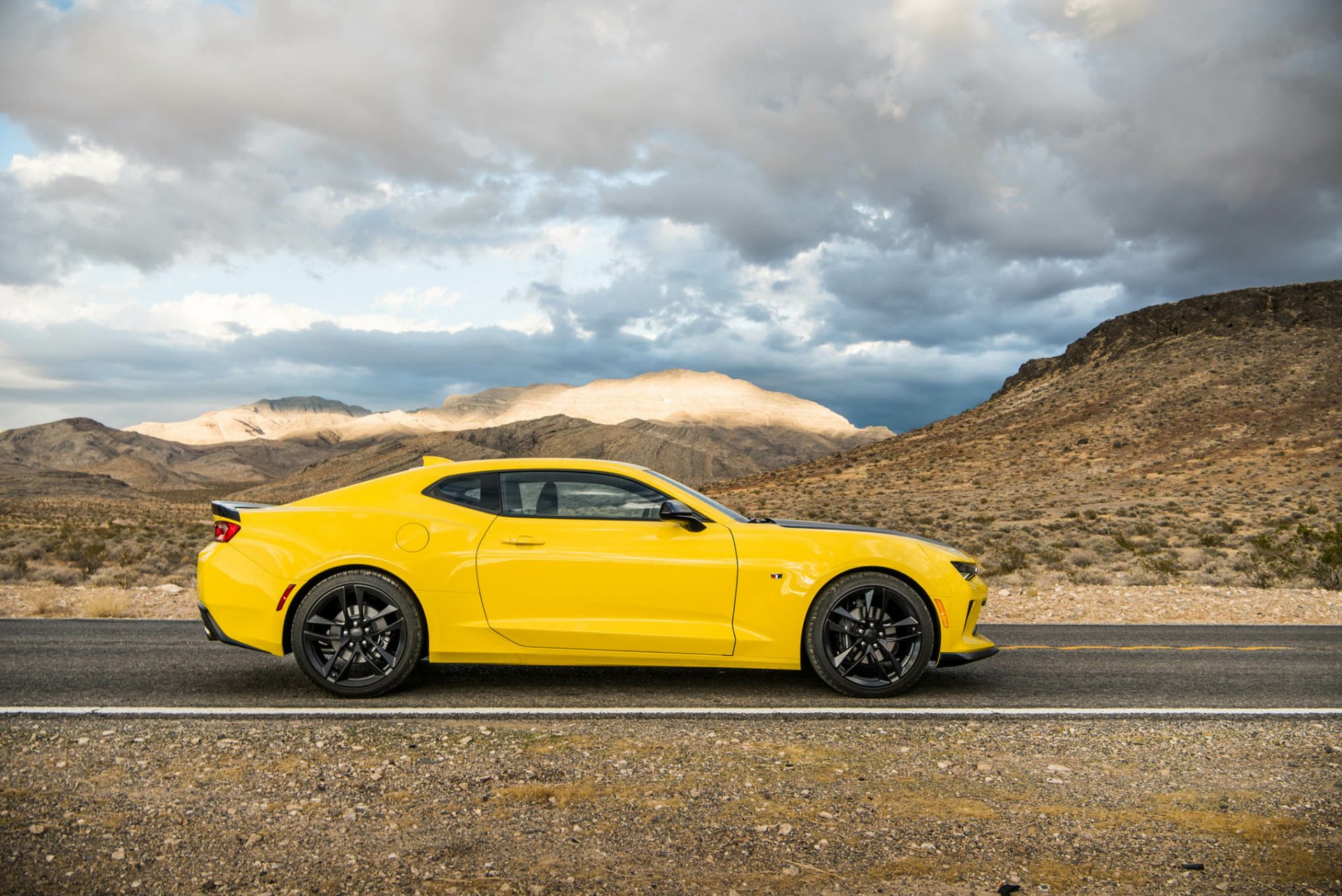 HD desktop wallpaper featuring a bright yellow Chevrolet Camaro 1LE muscle car parked on a desert road under a dramatic cloudy sky.
