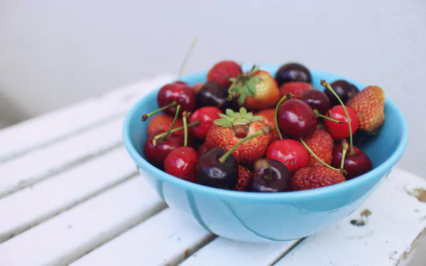 A 4K Ultra HD image of a blue bowl filled with fresh strawberries, cherries, and berries on a white wooden surface, showcasing vibrant food and fruit colors.