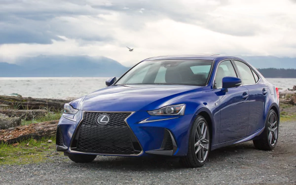 Blue Lexus IS car parked on a coastal gravel shore beneath dramatic clouds — 2K Quad HD PC desktop wallpaper background of a vehicle.