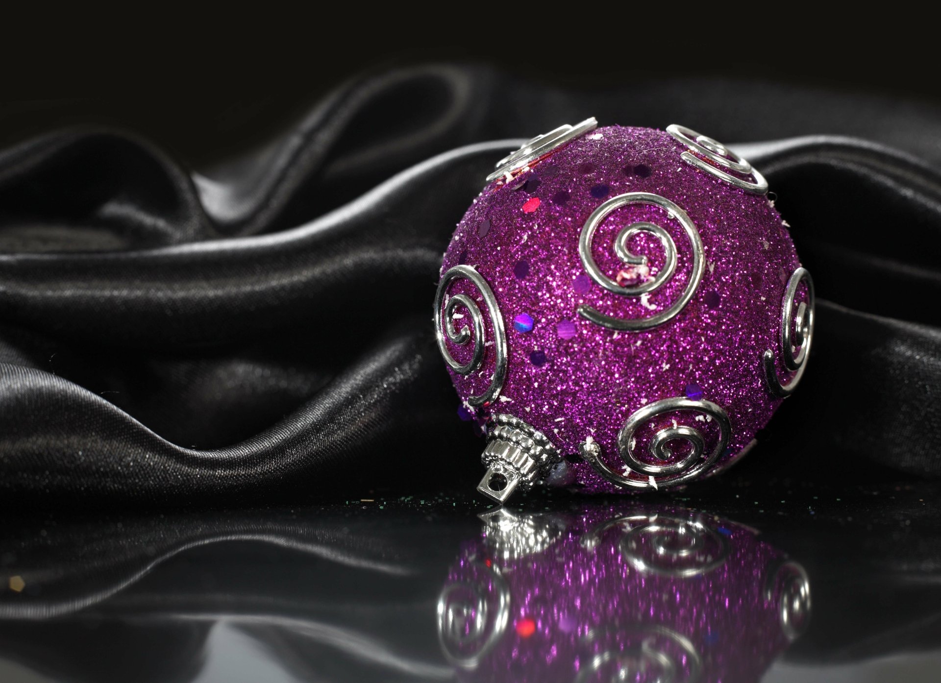 A sparkling purple Christmas ornament with silver swirls rests on a reflective surface against a smooth black fabric backdrop in 4K Ultra HD.